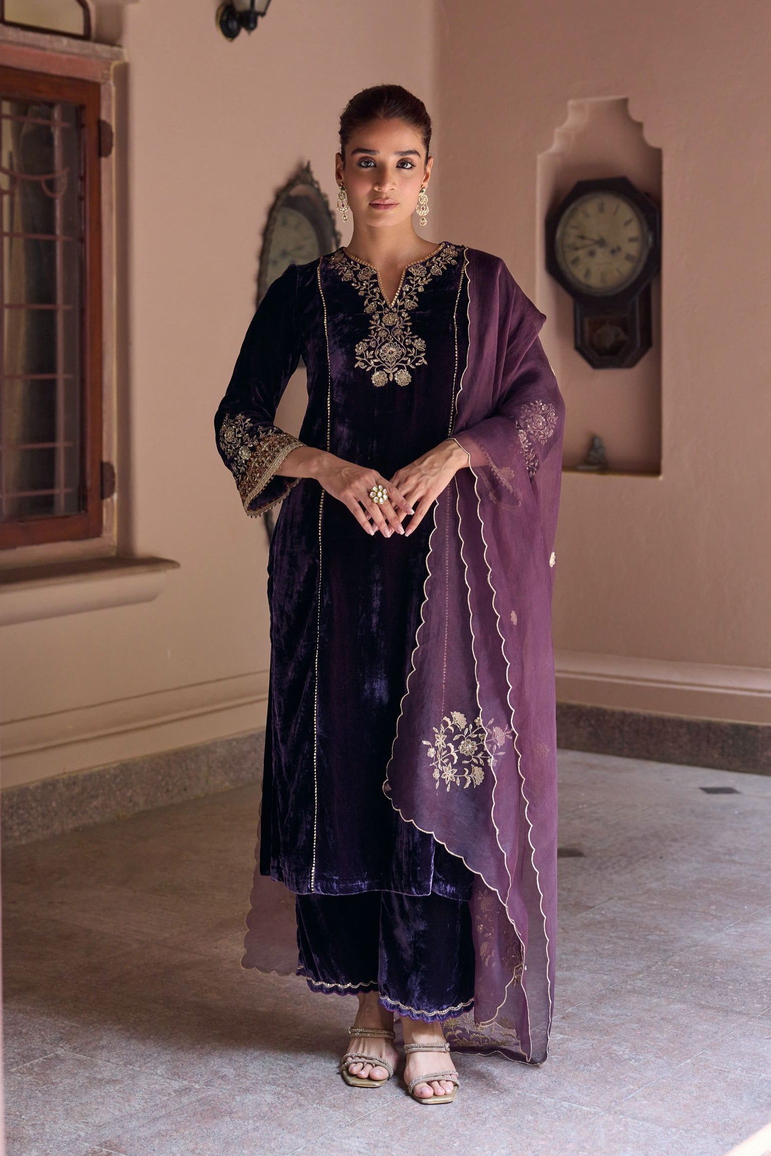 Woman in a dark purple traditional outfit standing in a room with a clock on the wall.